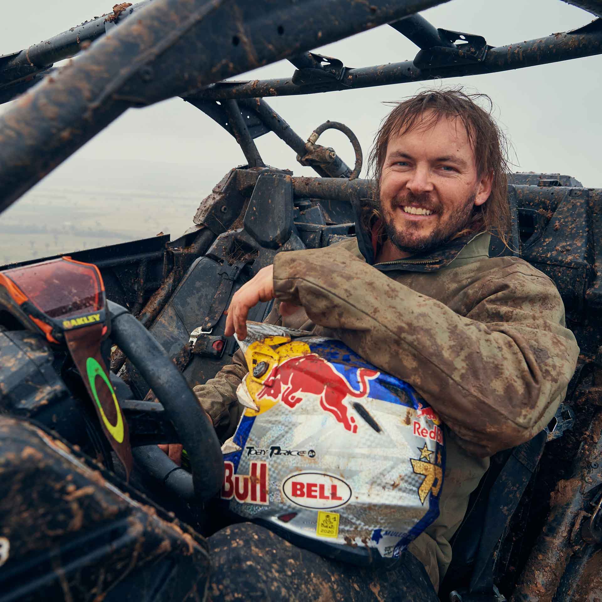 Toby Price smiling in a muddy Can-Am holding his helmet